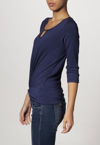 Tmavě modrý přiléhavý top s tříčtvrtečními rukávy, který má malý otvor u výstřihu a hladkou, jemnou texturu.