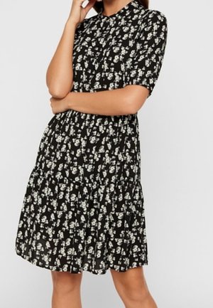 Kobieta w czarnej sukience do kolan w biały kwiatowy wzór, z krótkimi rękawami i kołnierzykiem, stoi z założonymi rękami.