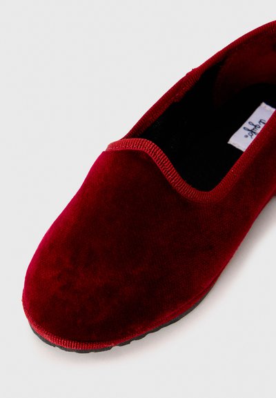 Červené zamatové slip-on topánky s jemnou textúrou, zaoblenou špičkou a užším elastickým lemovaním okolo otvoru. Čierna podrážka je viditeľná pod spodom.