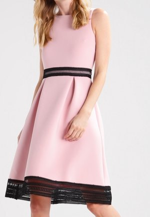 Vrouw draagt een mouwloze roze jurk met zwarte kanten rand bij taille en zoom tegen een effen lichte achtergrond.