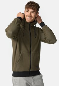 Olivgrüne Zip-Hoodie mit hohem Kragen, schwarzen Akzenten, verstellbaren Kordeln und frontalen Taschen. Der Stoff hat eine strukturierte Oberfläche.