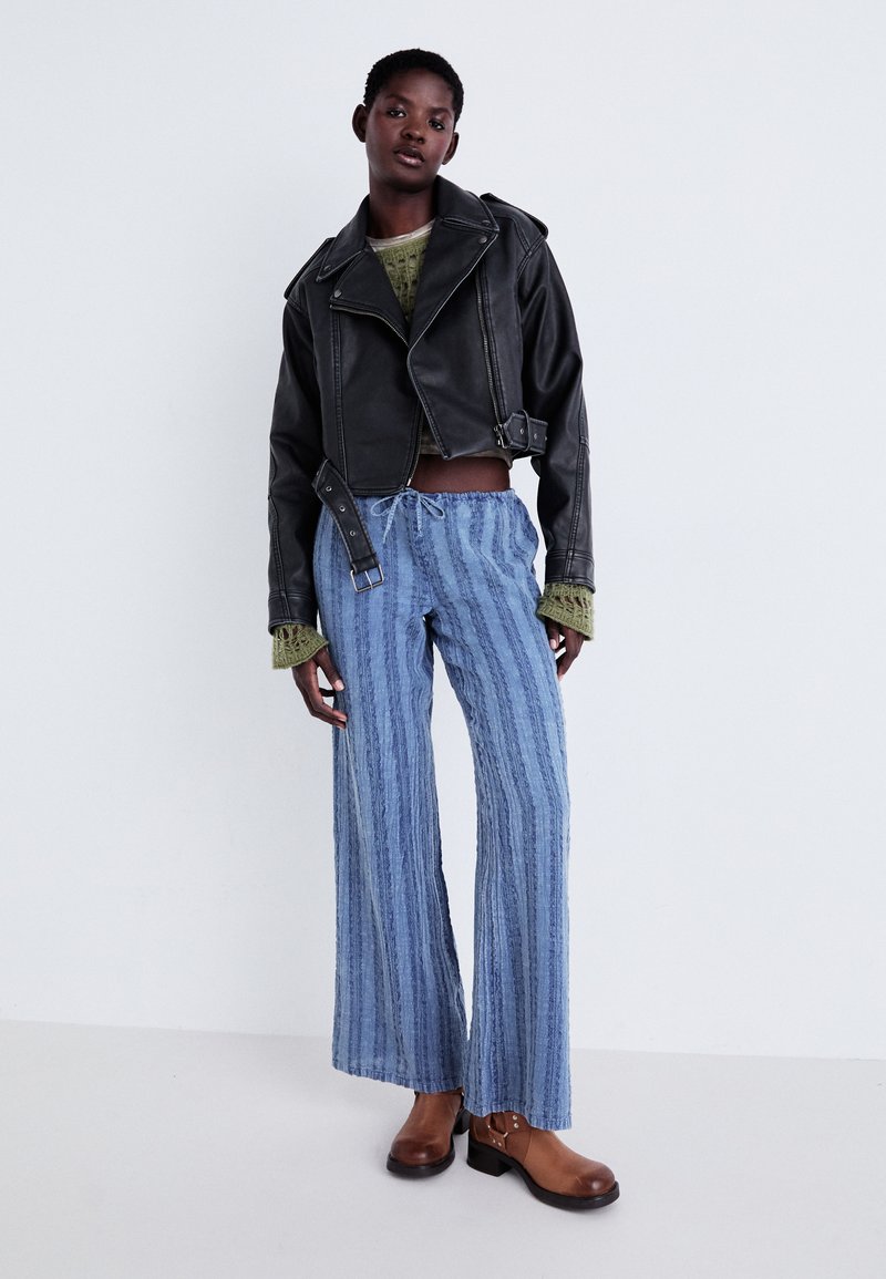 Veste en cuir noire courte, haut en tricot texturé vert, pantalon large à rayures bleu clair, et bottines à talons marron avec des accents métalliques.