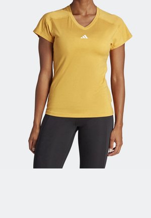 Vrouw draagt een geel Adidas sportshirt met korte mouwen en V-hals en zwarte legging, staand tegen een effen lichte achtergrond.