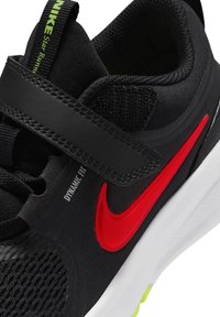 Μαύρο αθλητικό παπούτσι Nike με κόκκινο σήμα Swoosh, μαύρο ιμάντα με βέλκρο, λευκή σόλα και ετικέτα "Dynamic Fit" στο πλάι κοντά στα κορδόνια.