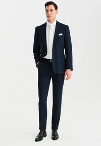 Granatowy garnitur z dopasowaną marynarką i pasującymi spodniami, biała koszula oraz biała muszka; czarne skórzane buty; poszetka w kieszeni piersiowej.