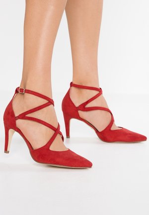 Talons hauts en daim rouge avec un bout pointu et des brides autour de la cheville, présentant des détails croisés et un talon fin. Texture lisse et sans motifs notables.