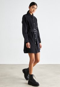 Černé džínové šatové šaty s knoflíkovou přední částí, dlouhými rukávy, páskem v oblasti pasu a náprsní kapsou. V kombinaci s černými platformovými botami na šněrování.