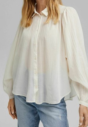 Kvinde iført en løs, langærmet hvid bluse med knapper, kombineret med lyseblå jeans mod en ens grå baggrund.