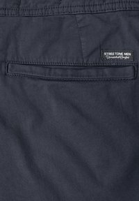 Poche arrière d’un pantalon bleu marine foncé avec une petite étiquette noire et blanche portant la mention « STREET ONE MEN Unmatched Comfort » près du bord supérieur de la poche.