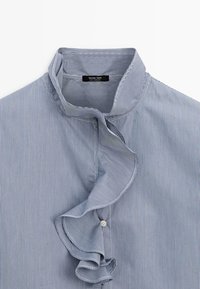 Chemise à fines rayures bleues et blanches avec un col montant et un détail volanté à l'avant autour de la patte de boutons.