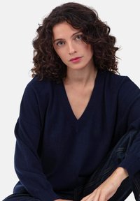 Pull en maille bleu marine à col en V avec manches longues et larges. Texture douce. Modèle assis, portant un jean, sur un fond blanc uni.