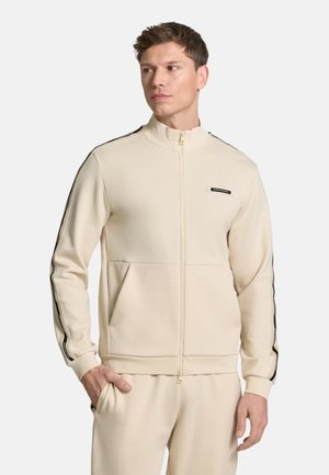 Uomo che indossa una giacca beige con zip della tuta con strisce nere sulle maniche e pantaloni abbinati, guarda verso sinistra con una mano in tasca.