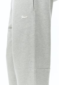 Lichtgrijze joggingsbroek gemaakt van een zachte stof, met een klein wit logo op de dij en een zijzak voor functionaliteit.