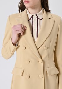 Blazer beige doppiopetto con rever a punta, tessuto testurizzato e tasche frontali. Presenta bottoni arrotondati e una camicia crema con colletto bordeaux.