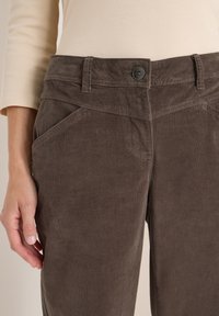 Pantaloni in velluto a coste marroni con una texture liscia, dotati di chiusura con bottone, tasche angolate e una vestibilità sartoriale attorno alla vita e ai fianchi.