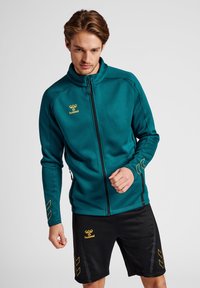 Teal ritsjack met een hoge kraag en een zwarte rits. Voorzien van een gouden logo en accenten. Wordt gedragen met zwarte shorts, ook met een gouden logo en ontwerp.