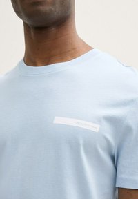 Svjetloplavi majica od mekog pamuka sa okruglim izrezom i bijelim etiketom koja se proteže preko prsa s natpisom "DENIM TOM TAILOR."