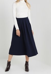 Jupe midi plissée bleu marine en tissu léger, associée à un haut ajusté à manches longues blanc et des bottes chevilles brillantes.