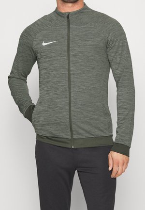 Homme portant une veste Nike gris-vert à manches longues zippée et un pantalon foncé, avec une main dans une poche et l'autre détendue.