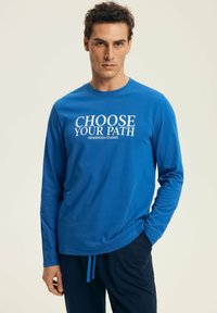 Man wearing a blue long-sleeve shirt with "CHOOSE YOUR PATH HENDERSON STUDIOS" text, paired with dark pants, standing against a plain background.