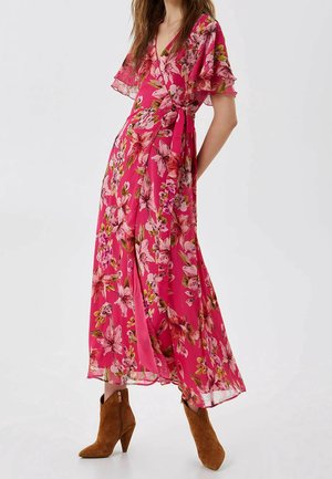 Femme portant une robe portefeuille fleurie rose avec des manches volantées et des bottines en daim marron, debout devant un fond uni.