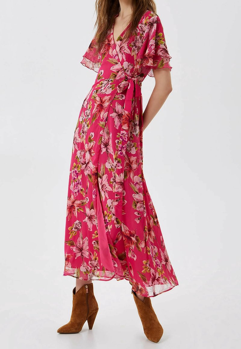 Femme portant une robe portefeuille fleurie rose avec des manches volantées et des bottines en daim marron, debout devant un fond uni.