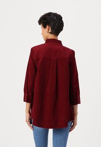 Burgunderfarget kordfløyelskjorte med lange ermer og knappemansjetter, med en bakgammel og buet hem, sammen med lyseblå jeans.