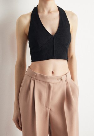 Woman wearing sleeveless black cropped halter top and high-waisted beige pleated trousers against plain white background.