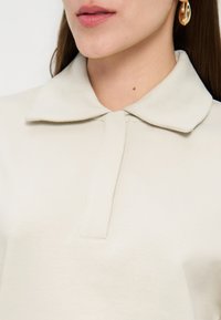 Tuvplāns sievietei, kura valkā krēmkrāsas kreklu ar apkakli un zelta stīpu auskarus, lūpas viegli atvērtas, ar gariem brūniem matiem.