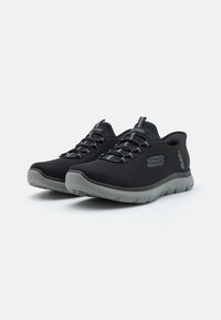 Pantofi sport negri din plasă, cu un superior texturat, șireturi elastice și o talpă gri cu un model în valuri pe partea inferioară.