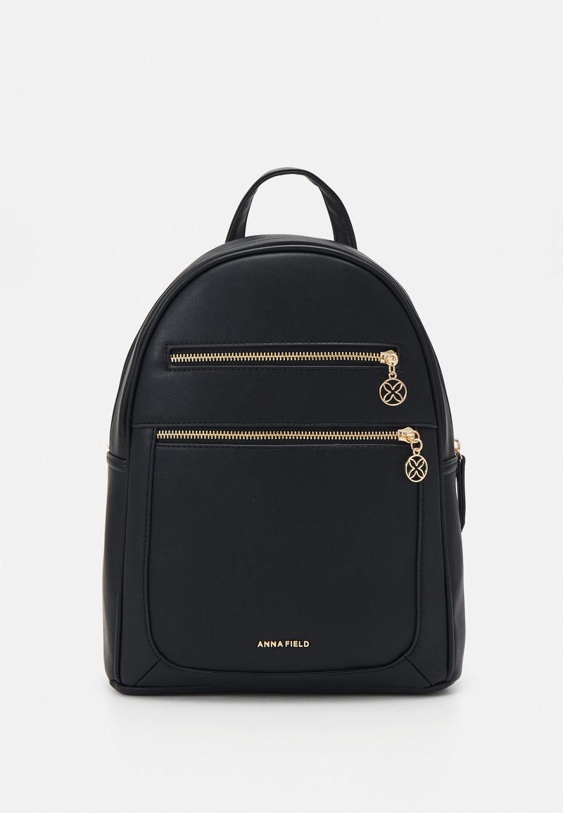 Mochila de imitación de cuero negra con forma redondeada, dos bolsillos frontales con cremallera y herrajes dorados, y un asa en la parte superior. Nombre de la marca "ANNA FIELD" en la parte frontal.