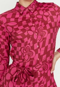 Košile s knoflíky v hluboké růžové barvě s výrazným květinovým a kostkovaným vzorem. Má límec a zvýrazněný pás na zavazování. Hladká textilie.