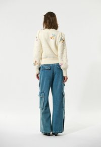 Pull en tricot crème avec texture en relief et broderies colorées ; associé à un pantalon cargo en denim bleu à jambe large avec poches latérales.