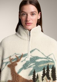 Biały polarowy pulower z wysokim kołnierzem, zamkiem na pół, z ozdobnym wzorem jelenia i gór w brązowym i niebieskim haftie.