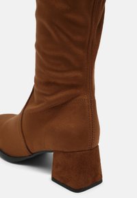 Botas de ante marrón hasta la mitad de la pantorrilla con tacón cuadrado y detalle de costura visible sobre un fondo blanco.