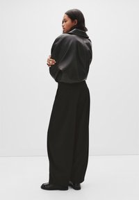 Črna usnjena oversize jakna z visokim ovratnikom, v kombinaciji z ohlapnimi črnimi hlačami in robustnimi črnimi čevlji. Gladka tekstura, minimalističen dizajn.