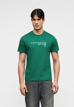 Ung mand iført en grøn Calvin Klein T-shirt med hvidt logo, kombineret med falmede mørkegrå jeans, stående mod en ensfarvet baggrund.