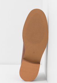 Chaussure plate marron avec une semelle en caoutchouc beige, fines rainures horizontales et marquage de pointure 37, semelle pressée verticalement contre un mur blanc.