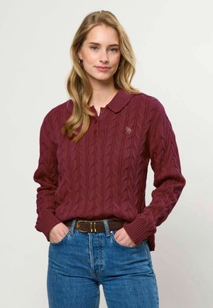 Frau mit langen blonden Haaren, die einen bordeauxfarbenen Zopfmuster-Pullover und blaue Jeans trägt, mit Händen in den Taschen vor einem einfarbigen hellen Hintergrund.