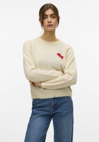 Maglione lavorato a maglia crema con scollo tondo, maniche lunghe e un motivo a cuore rosso sul petto. Abbinato a jeans in denim blu.