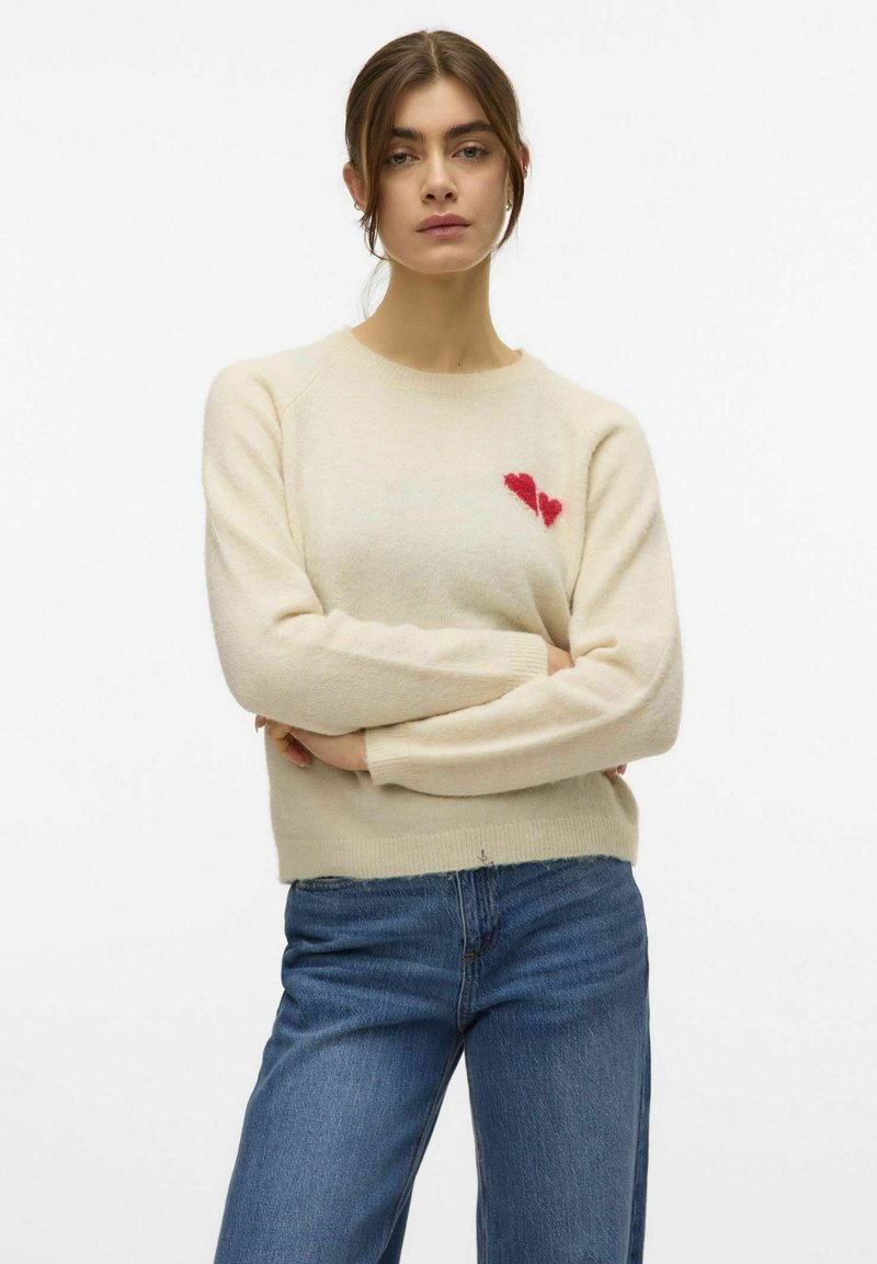 Maglione lavorato a maglia crema con scollo tondo, maniche lunghe e un motivo a cuore rosso sul petto. Abbinato a jeans in denim blu.