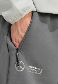 Grå joggingbukser med en sidelomme med lynlås, der har et Mercedes-logo og teksten "AMG PETRONAS Formula One Team". Glat tekstur og strømlinet design.