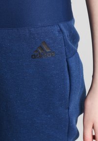 Detailní záběr na osobu v modrých sportovních kalhotách Adidas s viditelným logem u pravé kapsy a v modrém tričku, které k nim ladí.