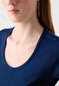 Navy blue fitted top with a scoop neckline and short sleeves. Text "calvin klein" printed subtly on the shoulder. Smooth fabric texture.