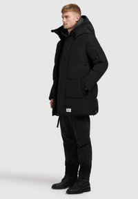 Svart vadderad parka med huva, med sidofickor, ribbad krage och justerbar nederkant. Bärs över en svart huvtröja och jeans.