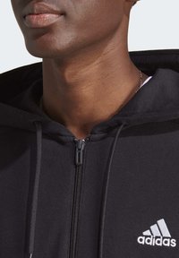Melnā kapuce ar rāvējslēdzēju un teksturētu apdari, ar sudraba Adidas logo kreisajā krūtīs un aukliņu detalizāciju uz kapuces.