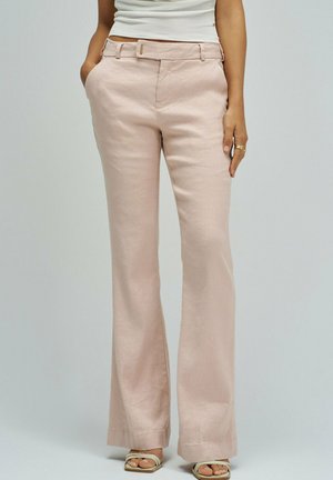Femme portant un pantalon large taille haute rose clair avec des poches et des talons ouverts blancs, debout devant un fond uni.