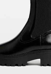 Stivale alla caviglia nero realizzato in materiale sintetico lucido con inserti in tessuto a coste. Presenta una suola spessa e robusta e un design elegante.