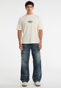 T-shirt oversize crema con grafica "ORIGINALS", abbinato a pantaloni cargo blu a vestibilità ampia, caratterizzati da più tasche e una texture sbiadita.