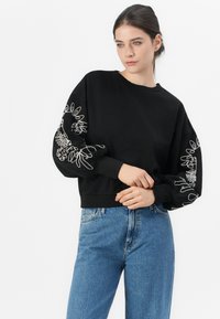 Kvinne i svart hettegenser med hvit abstrakt broderi på ermene og blå jeans, som står foran en enkel hvit bakgrunn.
